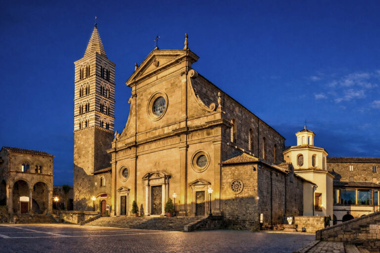 La Basilica di San Lorenzo a Viterbo illuminata all’ora blu, con la facciata in pietra chiara, il campanile romanico a fasce bianche e scure e la piazza lastricata nel centro storico della città laziale