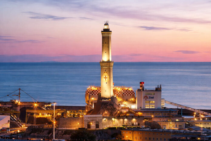 campanili-ditalia_liguria_genova_lanterna_redazione La Lanterna di Genova illuminata al tramonto domina il porto industriale ligure, con il faro storico al centro, il mare calmo sullo sfondo e le strutture portuali illuminate ai suoi piedi