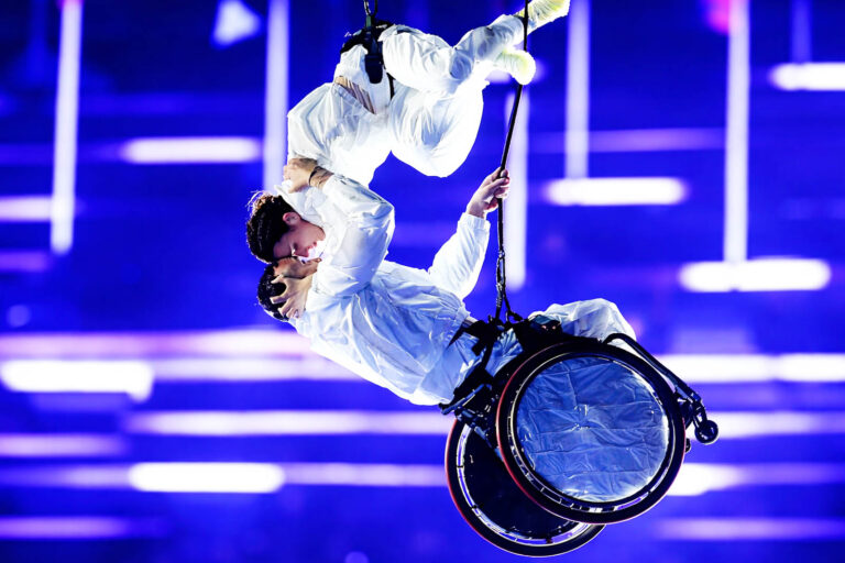 Artisti sospesi su cavi durante la cerimonia di chiusura delle Paralimpiadi Milano Cortina 2026, con una performer in carrozzina sollevata in aria in una coreografia luminosa su sfondo blu
