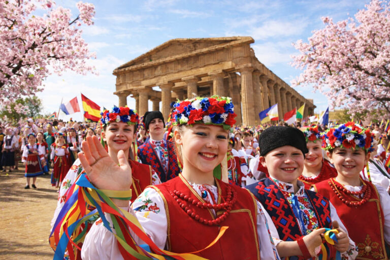 Bambini in costume tradizionale durante una festa internazionale davanti al tempio greco di Paestum, tra bandiere e alberi in fiore in primavera