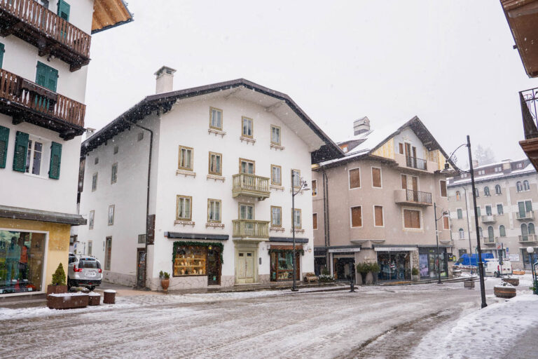 Centro storico di Cortina d’Ampezzo durante una nevicata invernale, con edifici tradizionali alpini, negozi al piano terra e strada coperta di neve nel cuore della località dolomitica