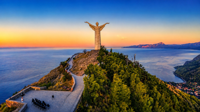 campaniliditalia_maratea-basilicata-internet Statua del Cristo Redentore di Maratea al tramonto, su promontorio panoramico affacciato sul mare Tirreno, con sentiero tortuoso, vegetazione verde e montagne sullo sfondo sotto un cielo limpido dai toni dorati e blu.