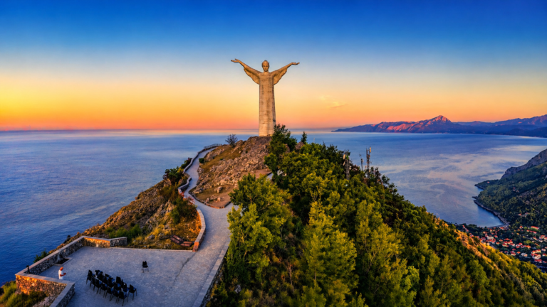 Statua del Cristo Redentore di Maratea al tramonto, su promontorio panoramico affacciato sul mare Tirreno, con sentiero tortuoso, vegetazione verde e montagne sullo sfondo sotto un cielo limpido dai toni dorati e blu.