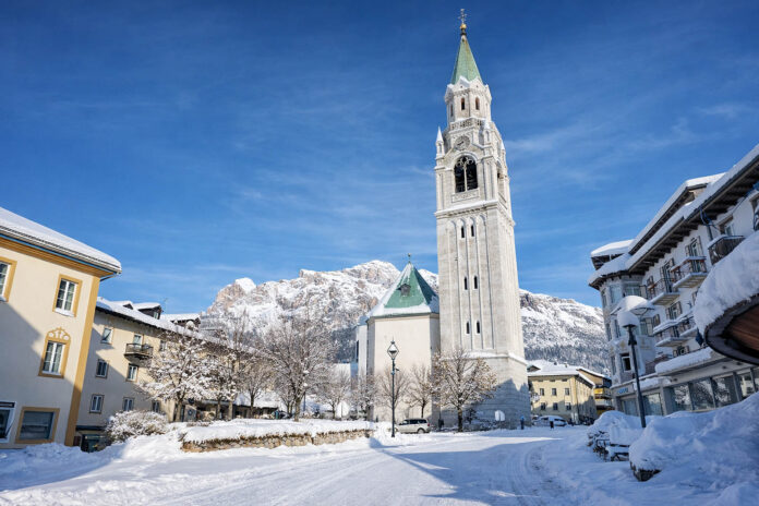 Campanile e chiesa di Cortina d’Ampezzo con la piazza coperta di neve e le Dolomiti innevate sullo sfondo in una giornata invernale